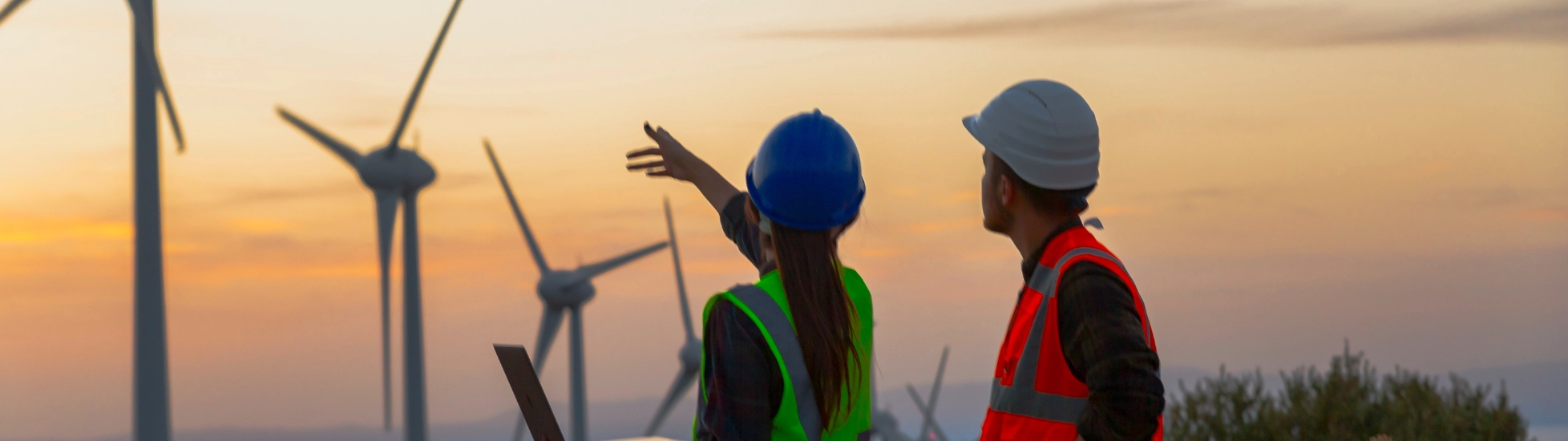 Two wind technicians (green vest/blue hard hat pointing with laptop; orange vest/white hard hat) overlook a field of large wind turbines at sunset.